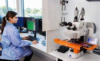 A female scientist in a blue lab coat sits at a computer beside a digital microscope scanning system.